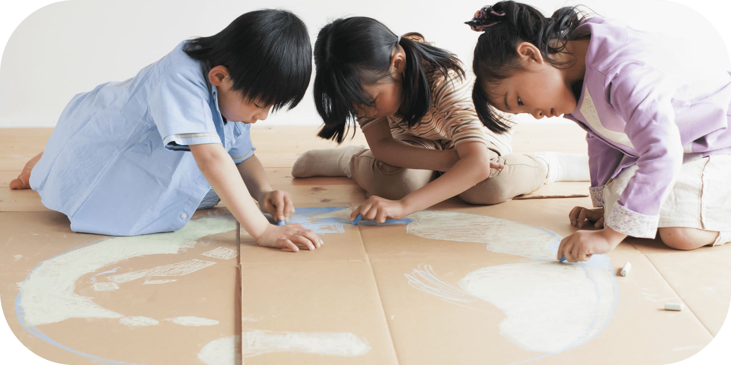 ３人の子供が大きな段ボールにチョークで絵を描いている。